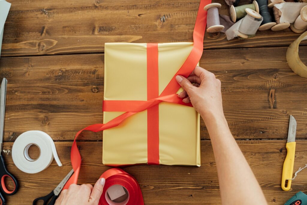 pexels-photo-5486890-5486890 Hands wrapping a gift with red ribbon on a wooden table, surrounded by crafting tools.