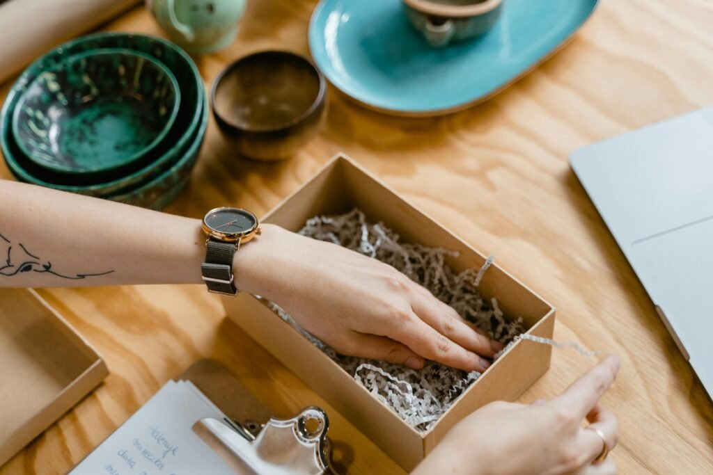 Close-up of a woman packing a cardboard box with shredded paper on a wooden desk.